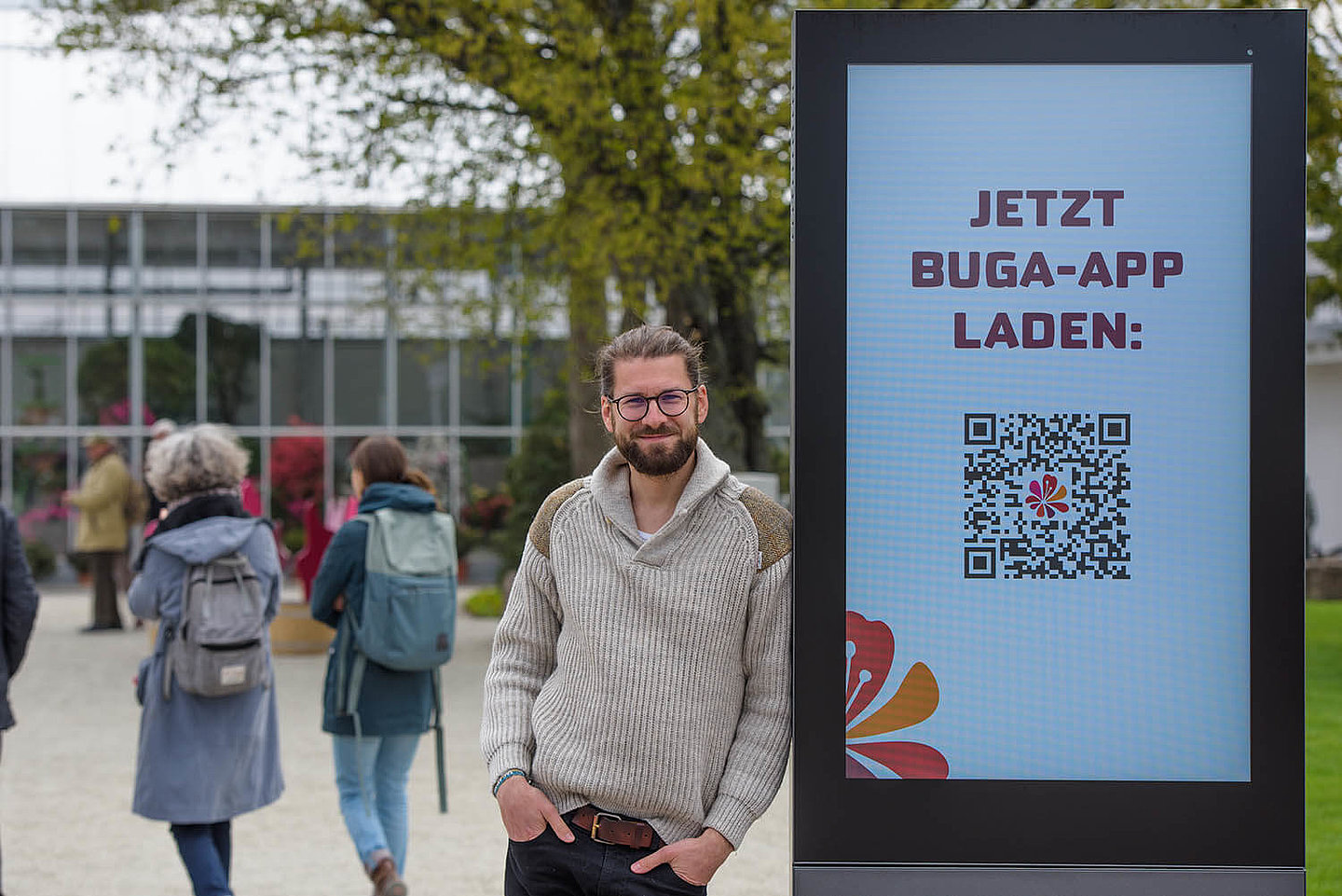 Eine Person, welche sich an eine Werbetafel der BUGA 2021 lehnt. Im Hintergrund befinden sich verschwommen gehende Menschen