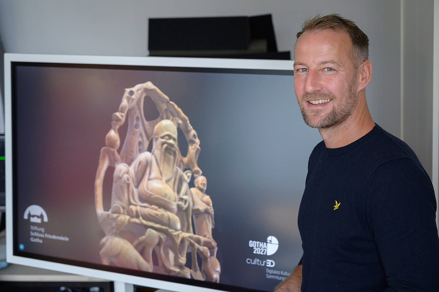 ein vor einem Monitor stehender mann schaut freundlich lächelnd in die Kamera