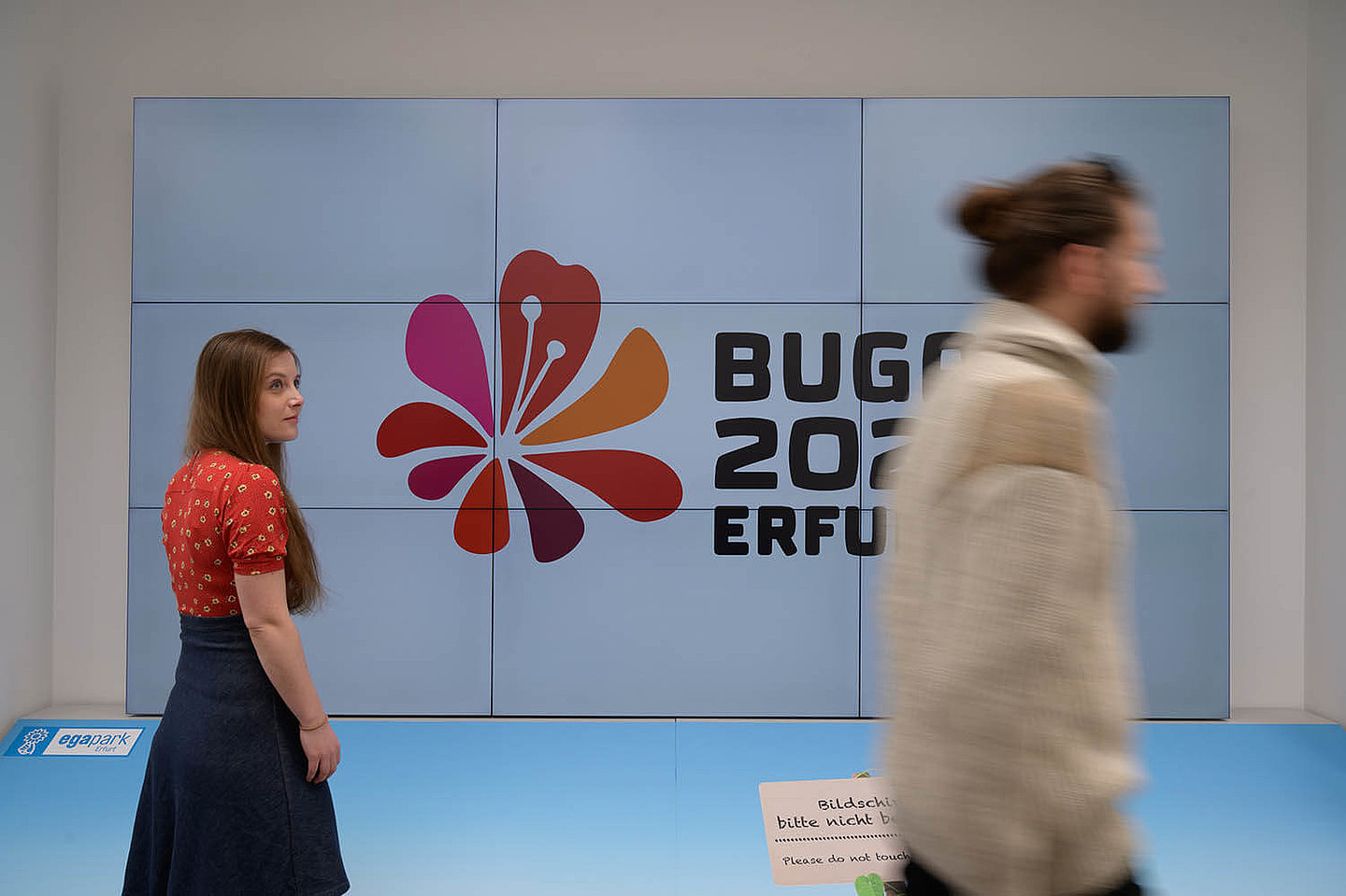 Zwei personen welche vor einem Bildschirm stehen, auf dem das Logo der BUGA 2021 abgebildet ist