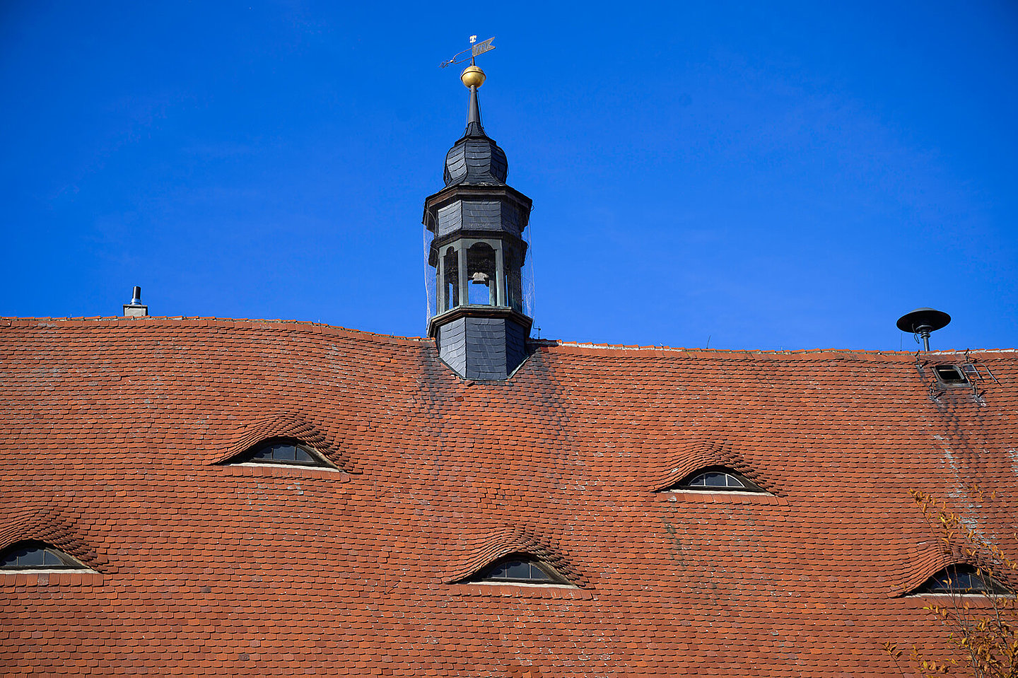 Eine Nahaufnahme des Glockenturm vom Buttstädter Rathaus. Dieser sitzt auf einem roten Dach mit kleinen Fenstern. In einer Ecke ist eine Alarmsirene montiert.