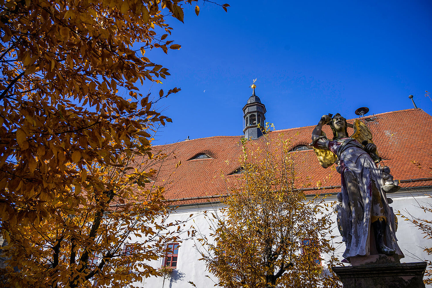 Im Vordergrund steht eine Steinskulptur eines Engels mit goldenen Flügeln. Im Hintergrund das Rathaus von Buttstätt