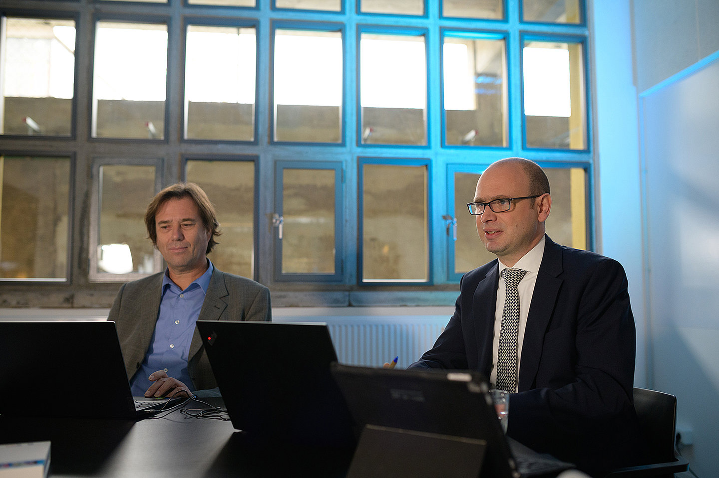 Dr. Laußmann und Herr Heinzel schauen auf Laptops. Sie sitzen in einem Studio der EFRE-Jahresveranstaltung 2021