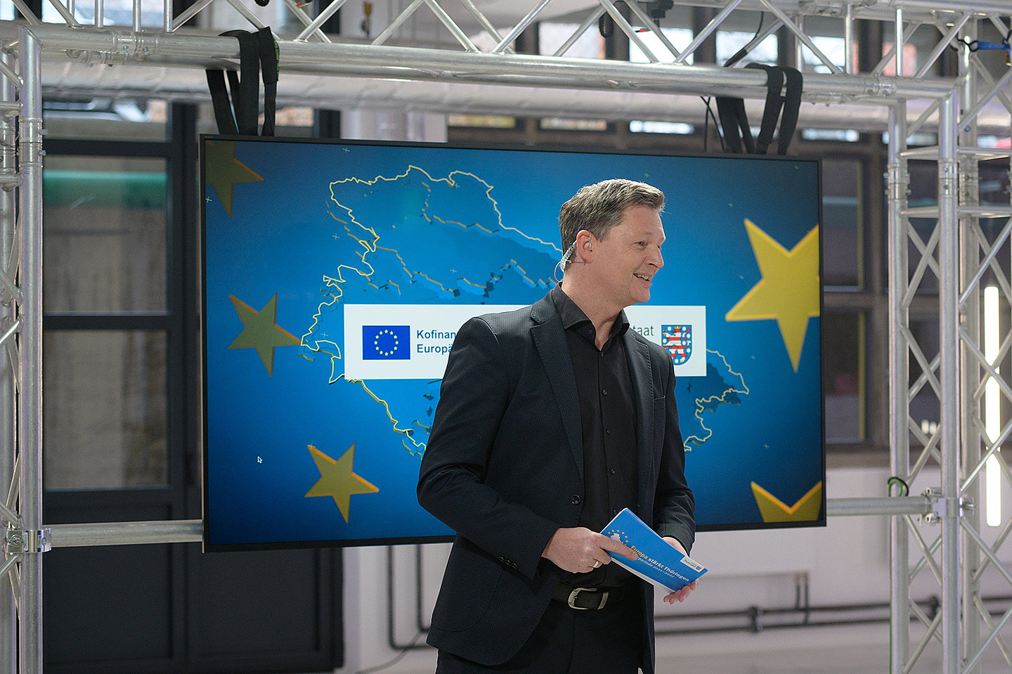 Der Moderator Andreas Fritsch steht lächelnd vor einem Monitor