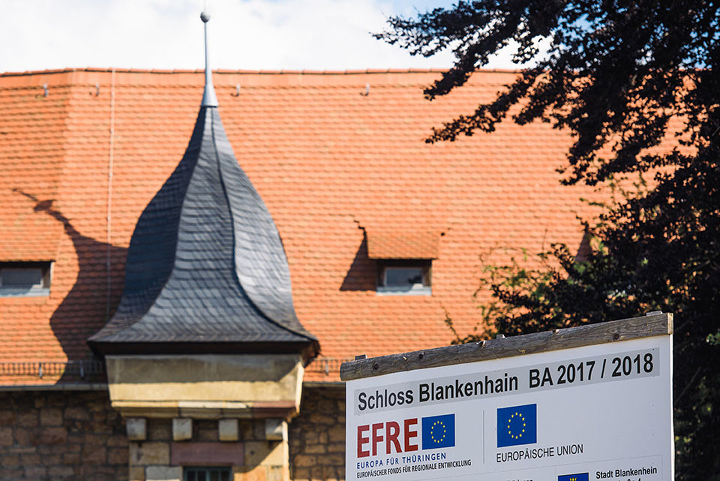 Im Vordergrund ist der Bauschild mit EFRE Logo zu sehen. Im Hintergrund ein Turm des Schloss Blankenhain und das Dach.