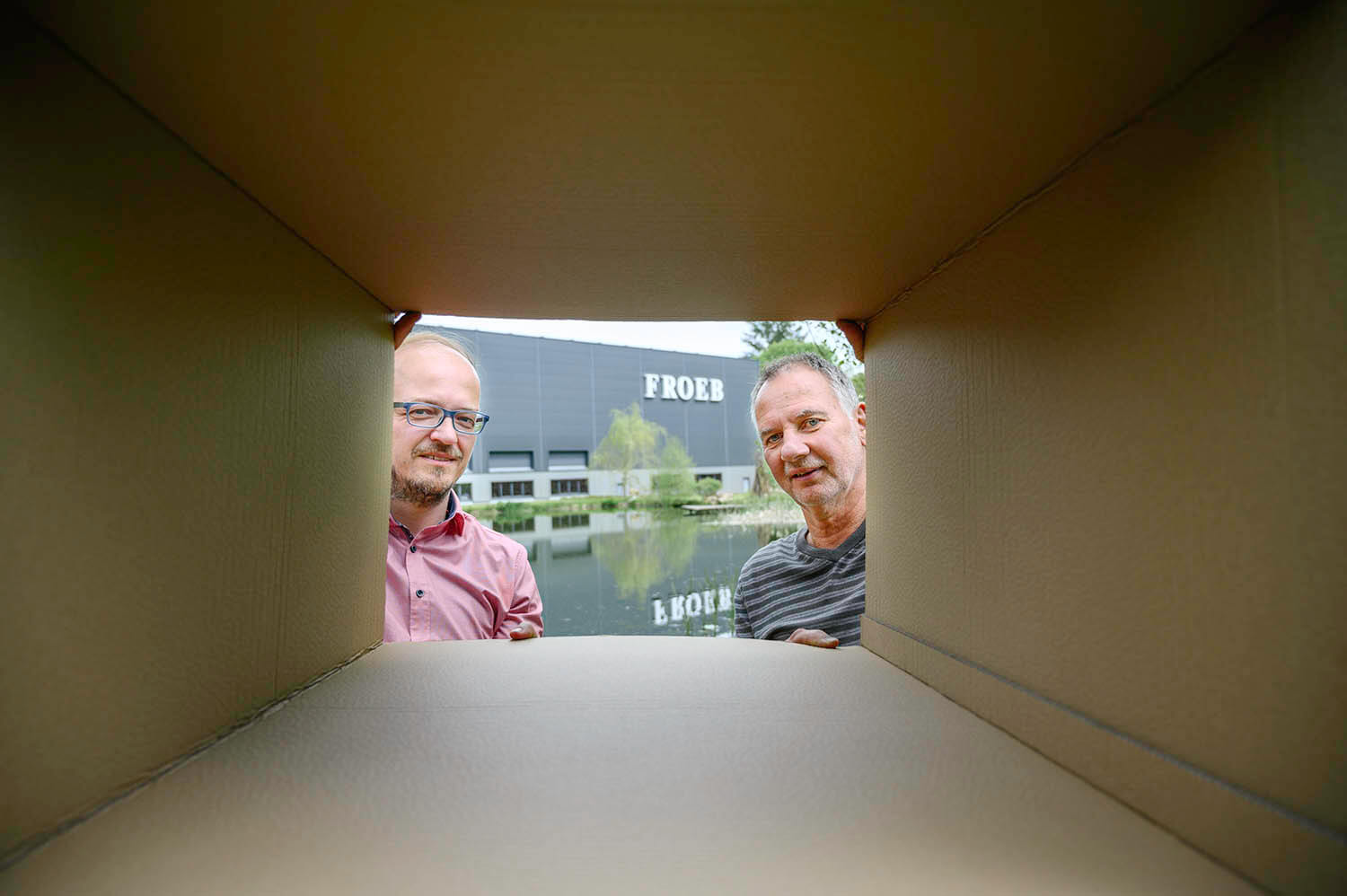 zwei Männer sehen durch einen Papkarton in die Kamera. Hinter ihnen befindet sich ein Haus der Froeb-Verpackungen GmbH